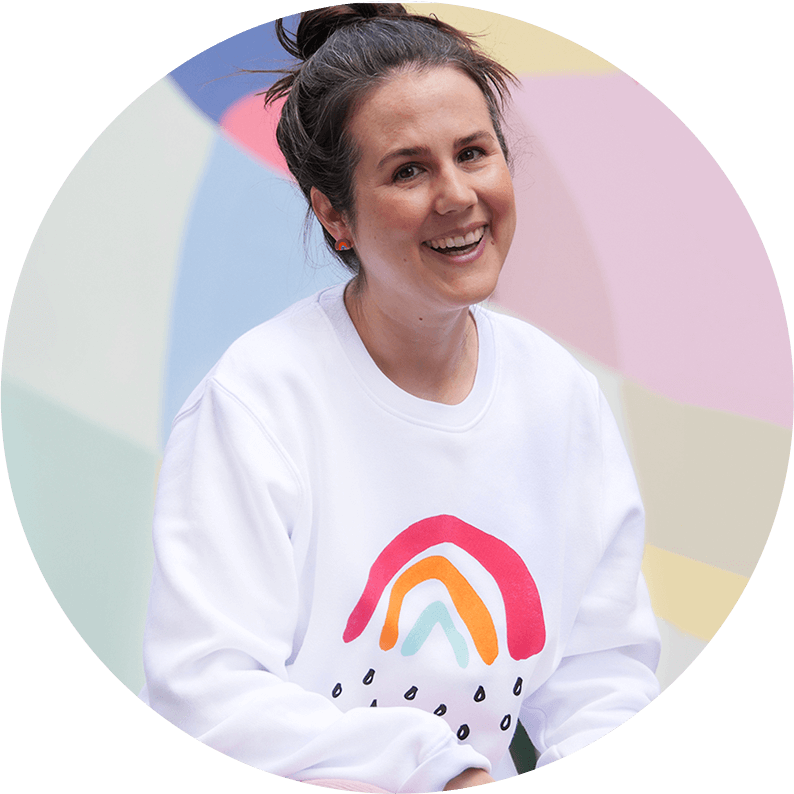 Sarah Gilgien, die Gründerin von Kleine Papeterie, ist in einem Pulli mit dem Kleinen Regenbogen vor einer bunten Wand zu sehen